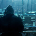 Hombre con capucha sentado frente a múltiples pantallas de computadora, trabajando en programación o hacking en un entorno oscuro y tecnológico.