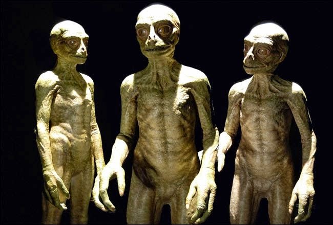 Tres figuras extraterrestres de aspecto humanoide, con piel verdosa y cabezas grandes, en un fondo oscuro. La imagen evoca temas de ciencia ficción y exploración espacial.