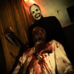 Hombre con máscara aterradora sosteniendo un hacha, de pie detrás de una figura ensangrentada, en un ambiente oscuro y escalofriante. Ideal para temáticas de terror y Halloween.