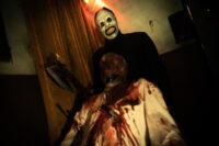 Hombre con máscara aterradora sosteniendo un hacha, de pie detrás de una figura ensangrentada, en un ambiente oscuro y escalofriante. Ideal para temáticas de terror y Halloween.