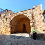 Arco de piedra en una antigua estructura histórica, con escaleras que conducen a un pasadizo, rodeado de paredes de piedra en un día soleado.