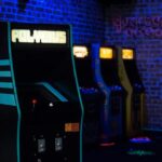 Máquina de arcade retro iluminada en un ambiente oscuro, con varias consolas de videojuegos clásicas en la fila. Ideal para amantes de los juegos vintage y la cultura gamer.