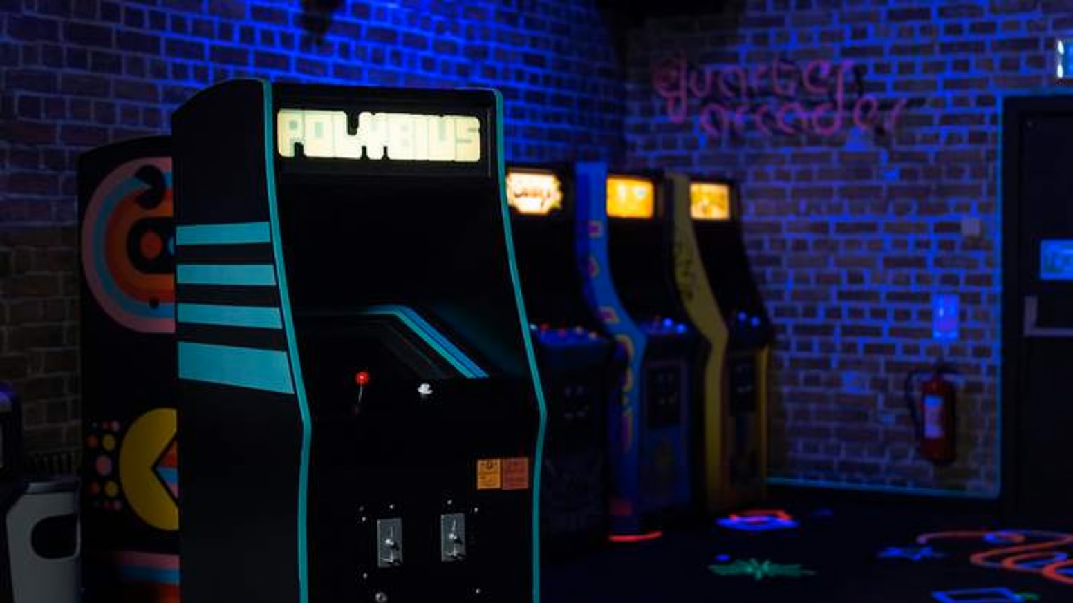 Máquina de arcade retro iluminada en un ambiente oscuro, con varias consolas de videojuegos clásicas en la fila. Ideal para amantes de los juegos vintage y la cultura gamer.