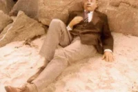 Hombre sentado en la playa, vestido con un traje oscuro y corbata, recostado sobre la arena y rodeado de rocas.