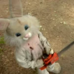 Muñeco de conejo con un hacha, en un entorno natural. El conejo tiene un aspecto siniestro, con manchas que simulan sangre en su traje. Ideal para contenido de terror o Halloween.