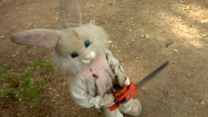 Muñeco de conejo con un hacha, en un entorno natural. El conejo tiene un aspecto siniestro, con manchas que simulan sangre en su traje. Ideal para contenido de terror o Halloween.