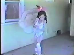 Niña pequeña vestida de bailarina con tutú blanco y diadema rosa, realizando un movimiento de danza en un entorno interior.