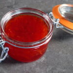 Frasco de mermelada roja casera con tapa de vidrio, sobre una superficie oscura. Ideal para acompañar tostadas y postres.