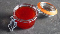 Frasco de mermelada roja casera con tapa de vidrio, sobre una superficie oscura. Ideal para acompañar tostadas y postres.
