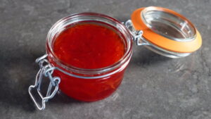 Frasco de mermelada roja casera con tapa de vidrio, sobre una superficie oscura. Ideal para acompañar tostadas y postres.