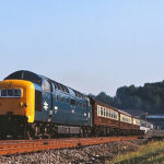 Locomotora de tren de pasajeros de color azul y amarillo en una vía férrea, con vagones de madera, rodeada de un paisaje verde y un cielo despejado.